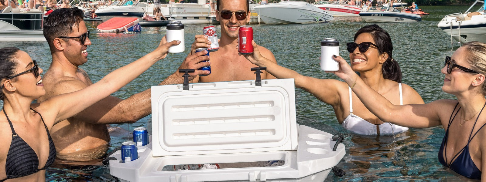 Group of five toasting drinks around a floating cooler in water with boats in the background.